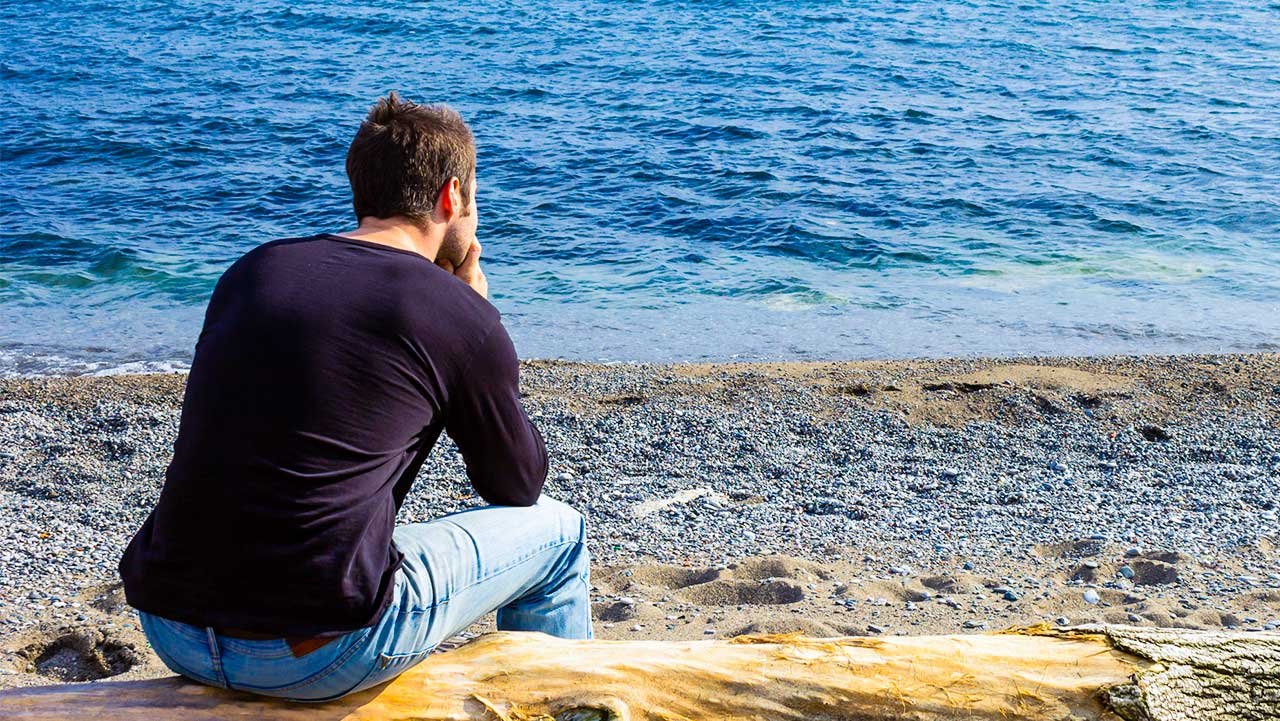 Mann sitzt nachdenklich auf einem Baumstamm am Meer