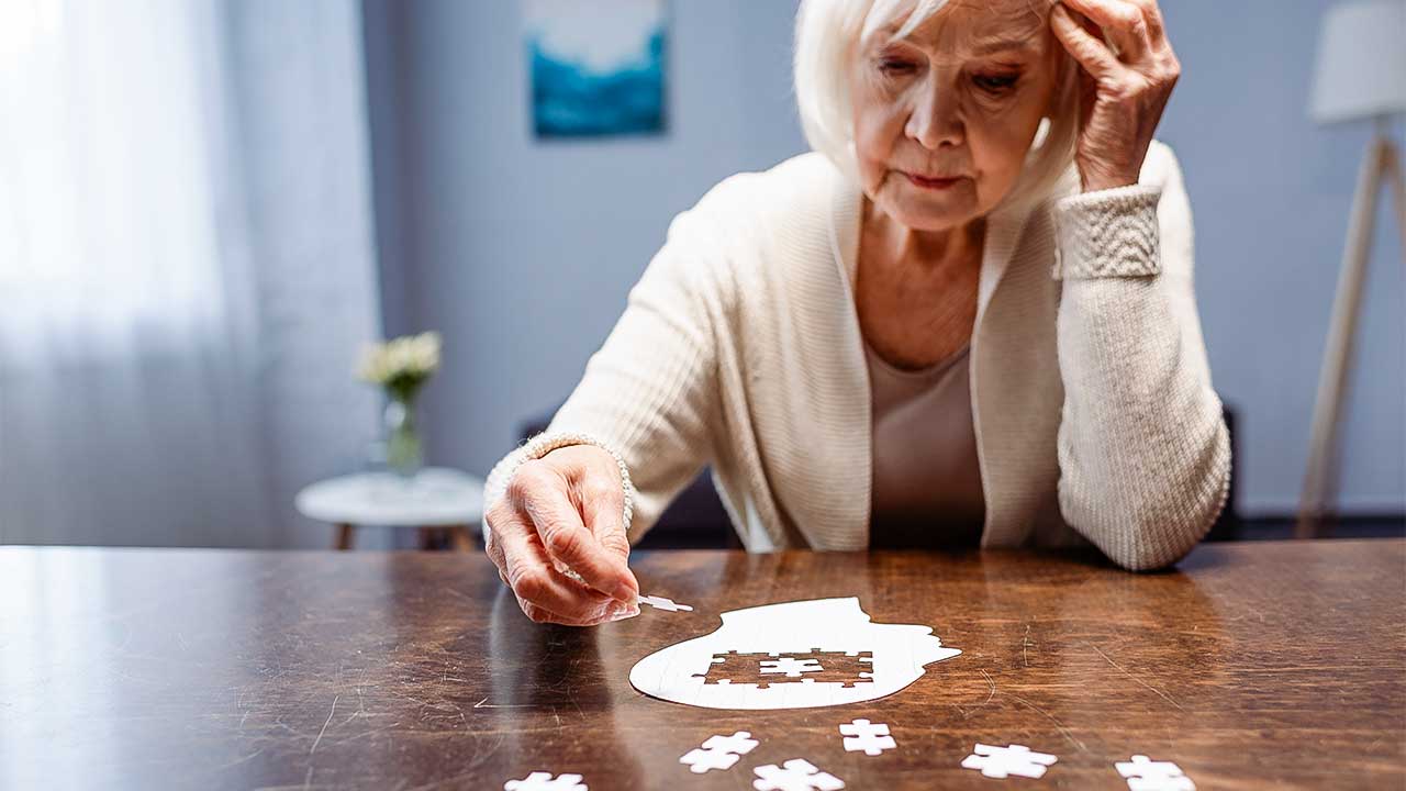 Seniorin legt auf einem Tisch ein Puzzle. Dieses hat die Form eines Kopfes mit einem Loch drin
